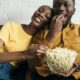 Casal negro feliz no sofá, comendo pipoca de uma tigela de vidro.