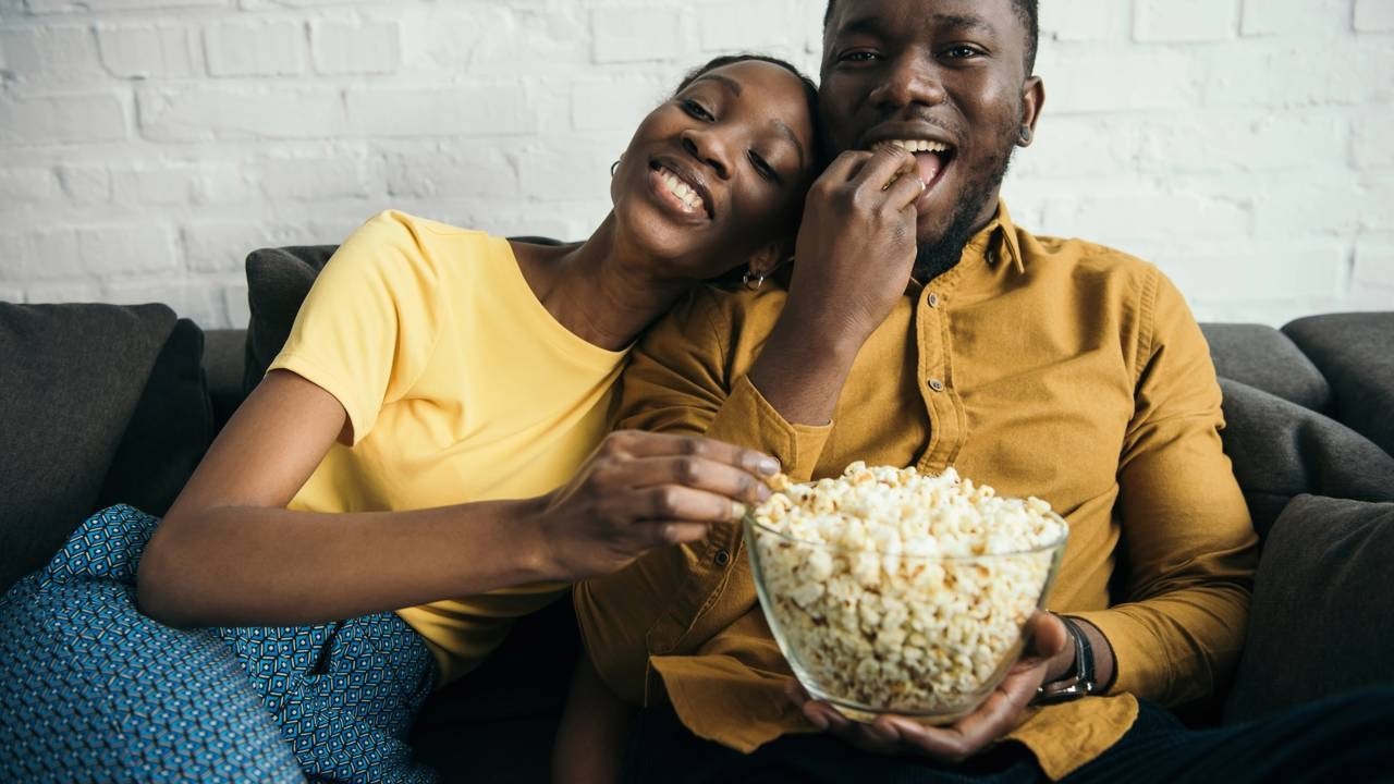 Casal negro feliz no sofá, comendo pipoca de uma tigela de vidro.