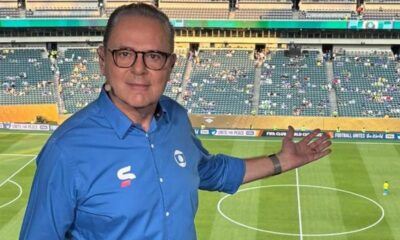 Luís Roberto de óculos e camisa azul da Globo gesticula em estádio de futebol.
