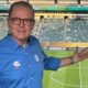 Luís Roberto de óculos e camisa azul da Globo gesticula em estádio de futebol.