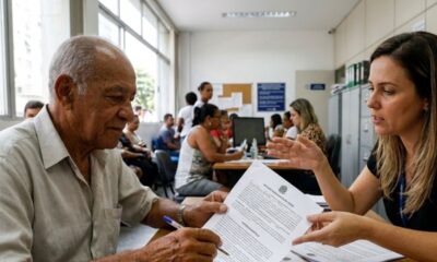 Homem idoso e mulher em mesa de cartório examinando documentos com uma caneta.