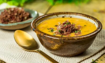 Tigela de caldo de mandioquinha cremoso com carne desfiada e salsinha, ao lado de colher dourada.