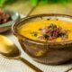 Tigela de caldo de mandioquinha cremoso com carne desfiada e salsinha, ao lado de colher dourada.