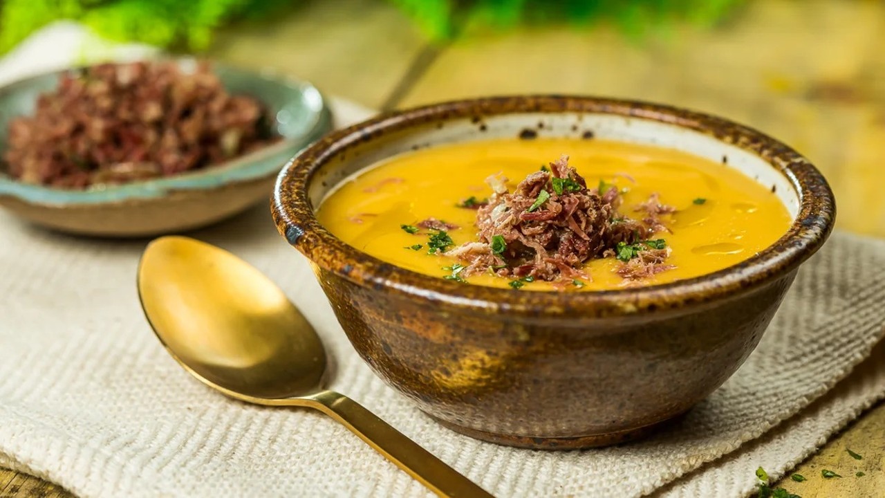 Tigela de caldo de mandioquinha cremoso com carne desfiada e salsinha, ao lado de colher dourada.