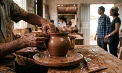 Mãos de artesão moldando um vaso de barro em roda de oleiro, com observadores ao fundo.