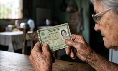 Mãos envelhecidas segurando uma carteira de identidade brasileira antiga com foto de jovem.
