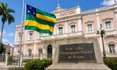 Monumento com a bandeira de Sergipe hasteada em frente ao Palácio-Museu Olímpio Campos, sob céu limpo, com placa em destaque escrita “8 de Julho – Emancipação Política de Sergipe”, em cenário realista que simboliza a autonomia e a identidade sergipana.