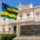 Monumento com a bandeira de Sergipe hasteada em frente ao Palácio-Museu Olímpio Campos, sob céu limpo, com placa em destaque escrita “8 de Julho – Emancipação Política de Sergipe”, em cenário realista que simboliza a autonomia e a identidade sergipana.