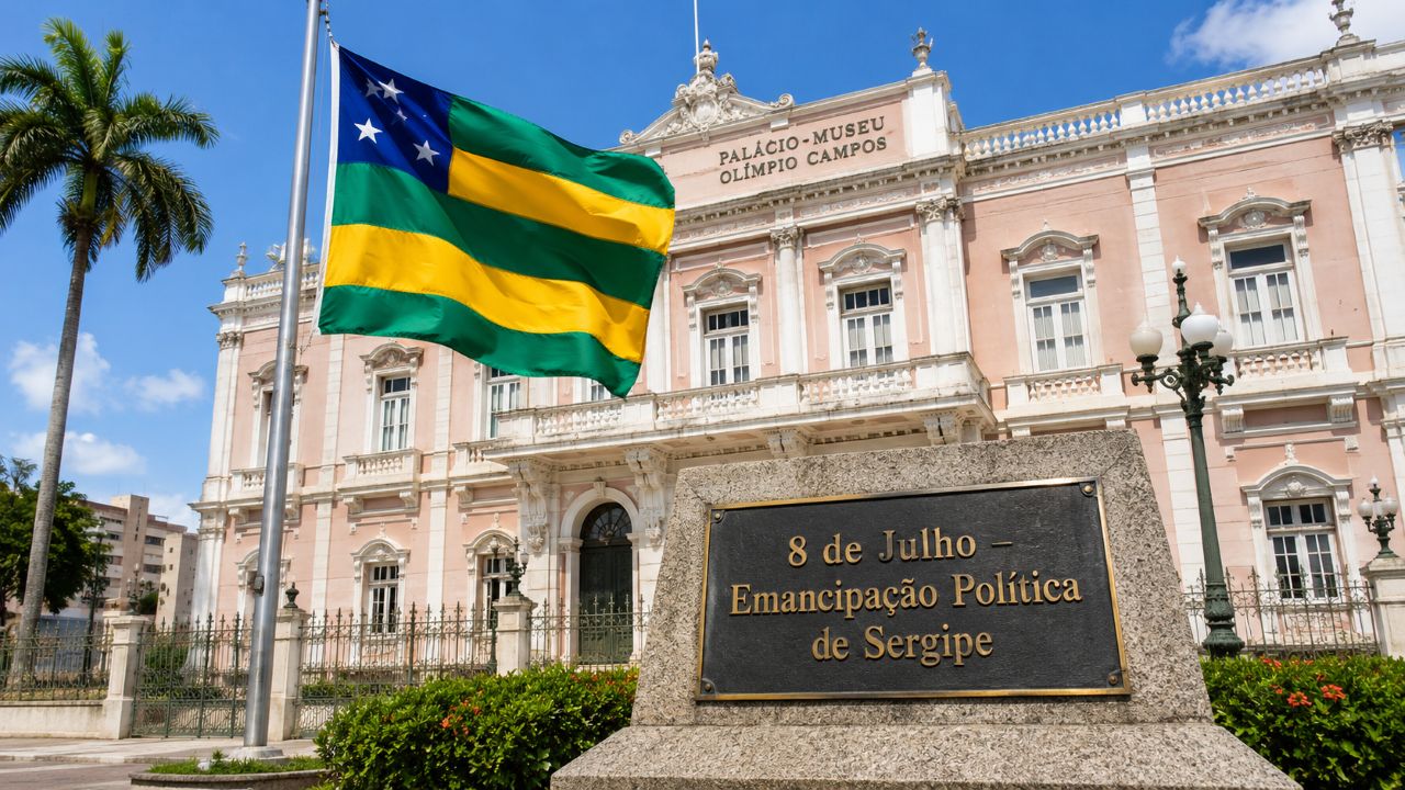 Monumento com a bandeira de Sergipe hasteada em frente ao Palácio-Museu Olímpio Campos, sob céu limpo, com placa em destaque escrita “8 de Julho – Emancipação Política de Sergipe”, em cenário realista que simboliza a autonomia e a identidade sergipana.