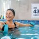 Pessoa praticando hidroginástica com halteres aquáticos em piscina coberta.