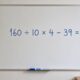 Quadro branco em sala de estudo limpa com o cálculo “160 ÷ 10 × 4 − 39 = ?” escrito em azul e um marcador sobre a bandeja inferior.