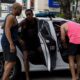 Três homens perto de carro policial branco com "DEA" e "67-2612" na porta aberta