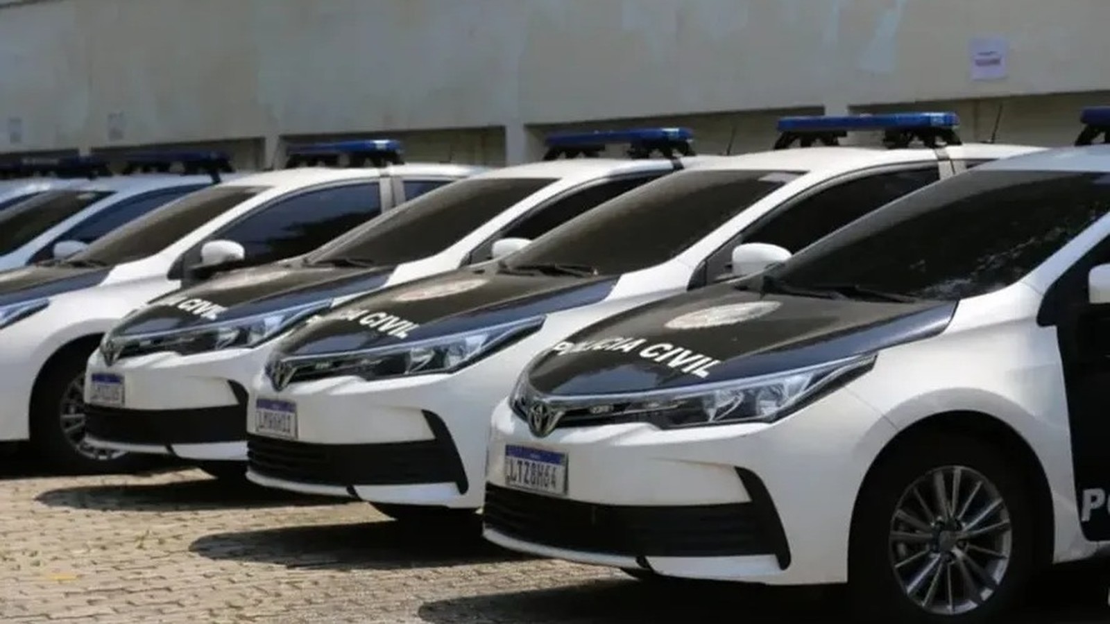 Viaturas da Polícia Civil, brancas e pretas, com luzes azuis no teto, estacionadas