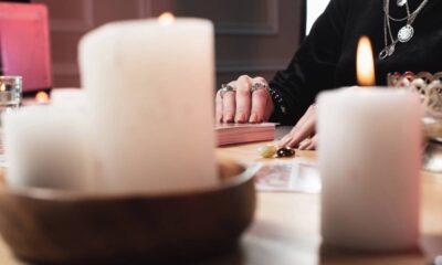 Mãos sobre baralho de cartas com anéis, velas acesas e pedras em uma mesa de madeira.