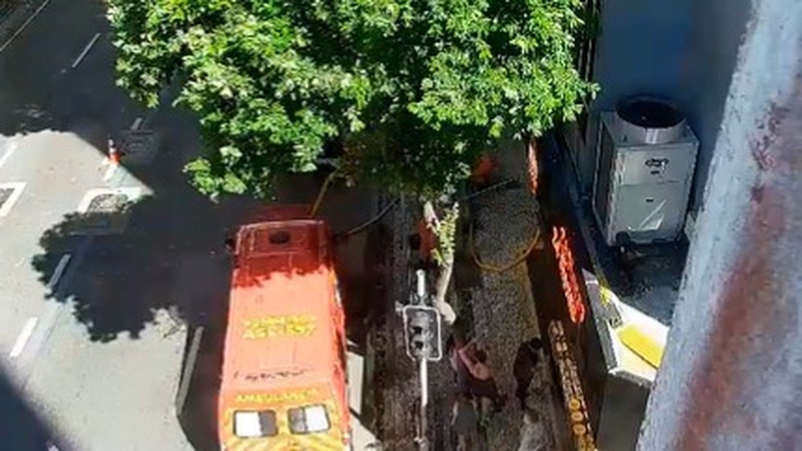 Ambulância laranja ‘AMBULÂNCIA ASSIST’ na rua, pessoas, árvore, unidade de ar condicionado