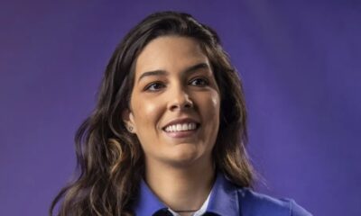 Mulher sorridente com cabelo escuro e camisa azul em fundo roxo