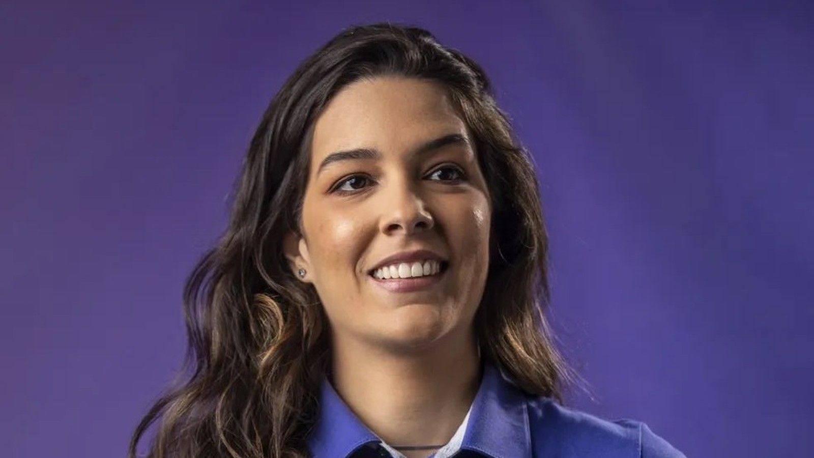 Mulher sorridente com cabelo escuro e camisa azul em fundo roxo