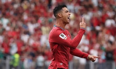 Cristiano Ronaldo celebra em campo com camisa vermelha, apontando os dedos para cima e à frente