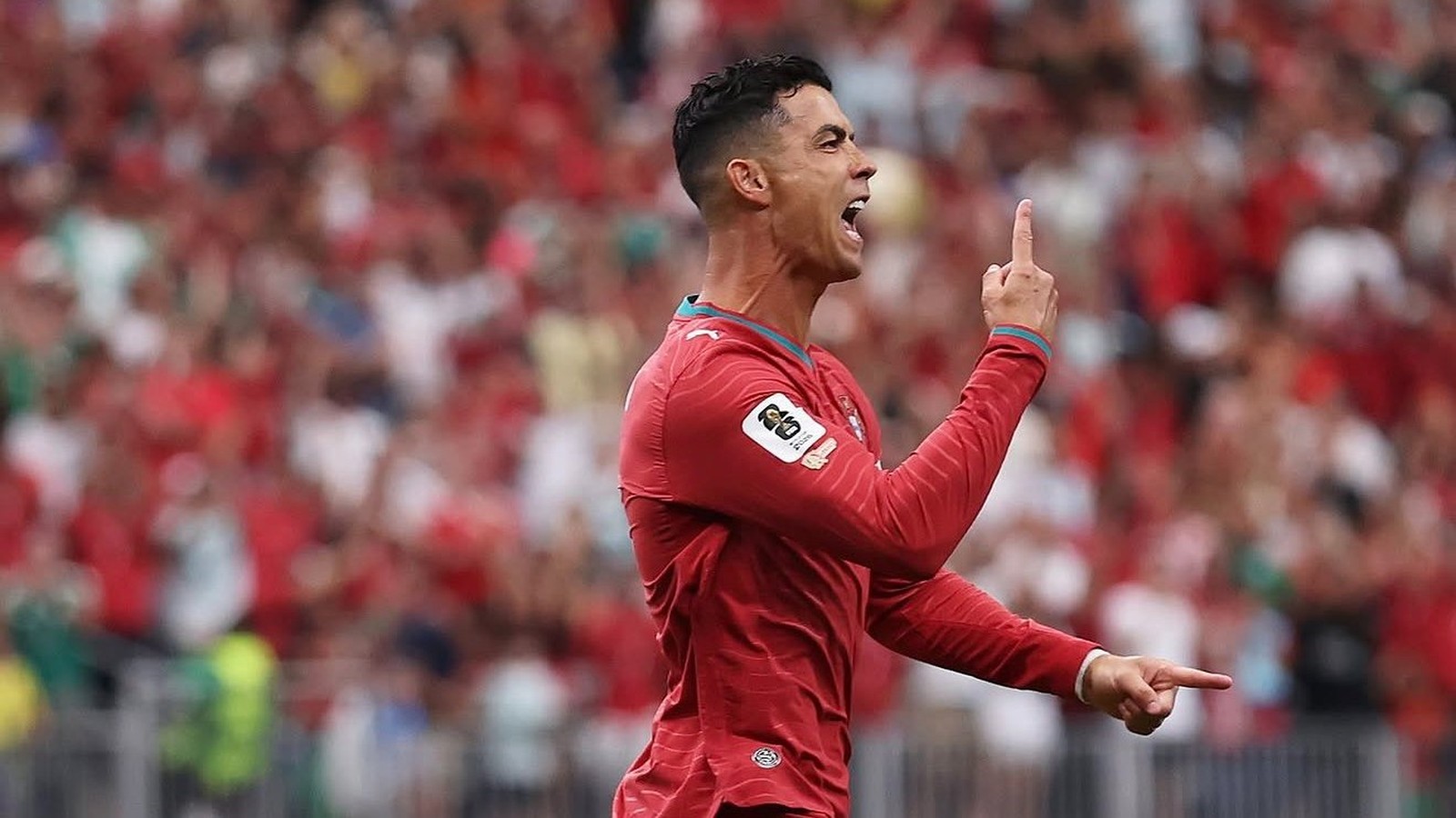 Cristiano Ronaldo celebra em campo com camisa vermelha, apontando os dedos para cima e à frente