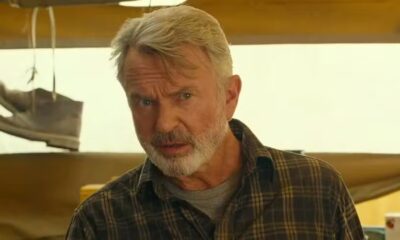 Sam Neill com cabelo grisalho e barba, camisa xadrez, olhando fixamente, com um sapato pendurado