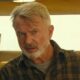 Sam Neill com cabelo grisalho e barba, camisa xadrez, olhando fixamente, com um sapato pendurado
