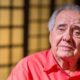 Luiz Carlos Barreto, homem idoso com cabelo grisalho e camisa vermelha, olhando pensativo