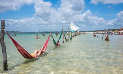 Pessoas relaxando em redes vermelha e verde na água rasa da praia sob céu azul.