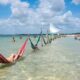 Pessoas relaxando em redes vermelha e verde na água rasa da praia sob céu azul.