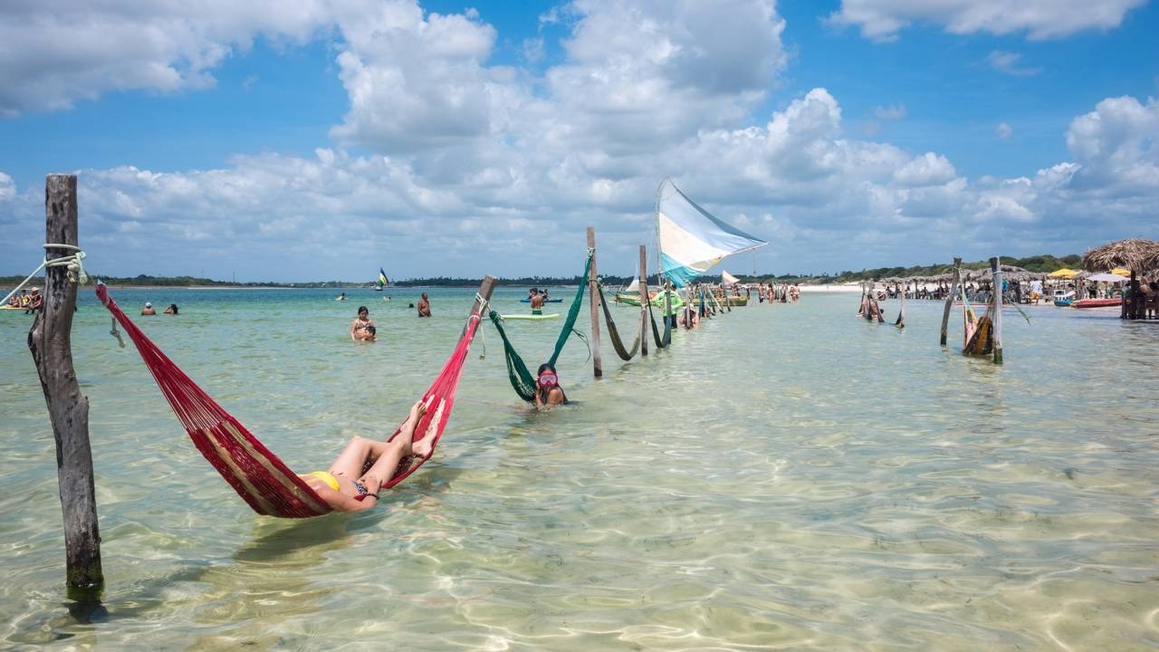 Pessoas relaxando em redes vermelha e verde na água rasa da praia sob céu azul.