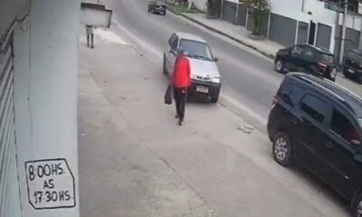Homem em vermelho na calçada, carro prateado ao lado. Poste com sinal "8:00HS. AS 17:30HS".