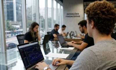 Grupo de pessoas trabalha em laptops, um com gráficos financeiros, em escritório com 'Inovações Financeiras' na parede