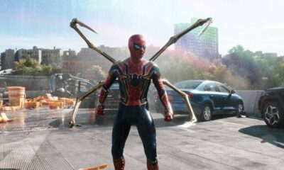 Homem-Aranha com traje vermelho e dourado, braços mecânicos, em rodovia destruída e chuvosa.