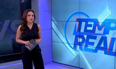 Apresentadora Márcia Dantas em estúdio, roupa preta, tablet nas mãos, tela "Tempo Real" ao fundo