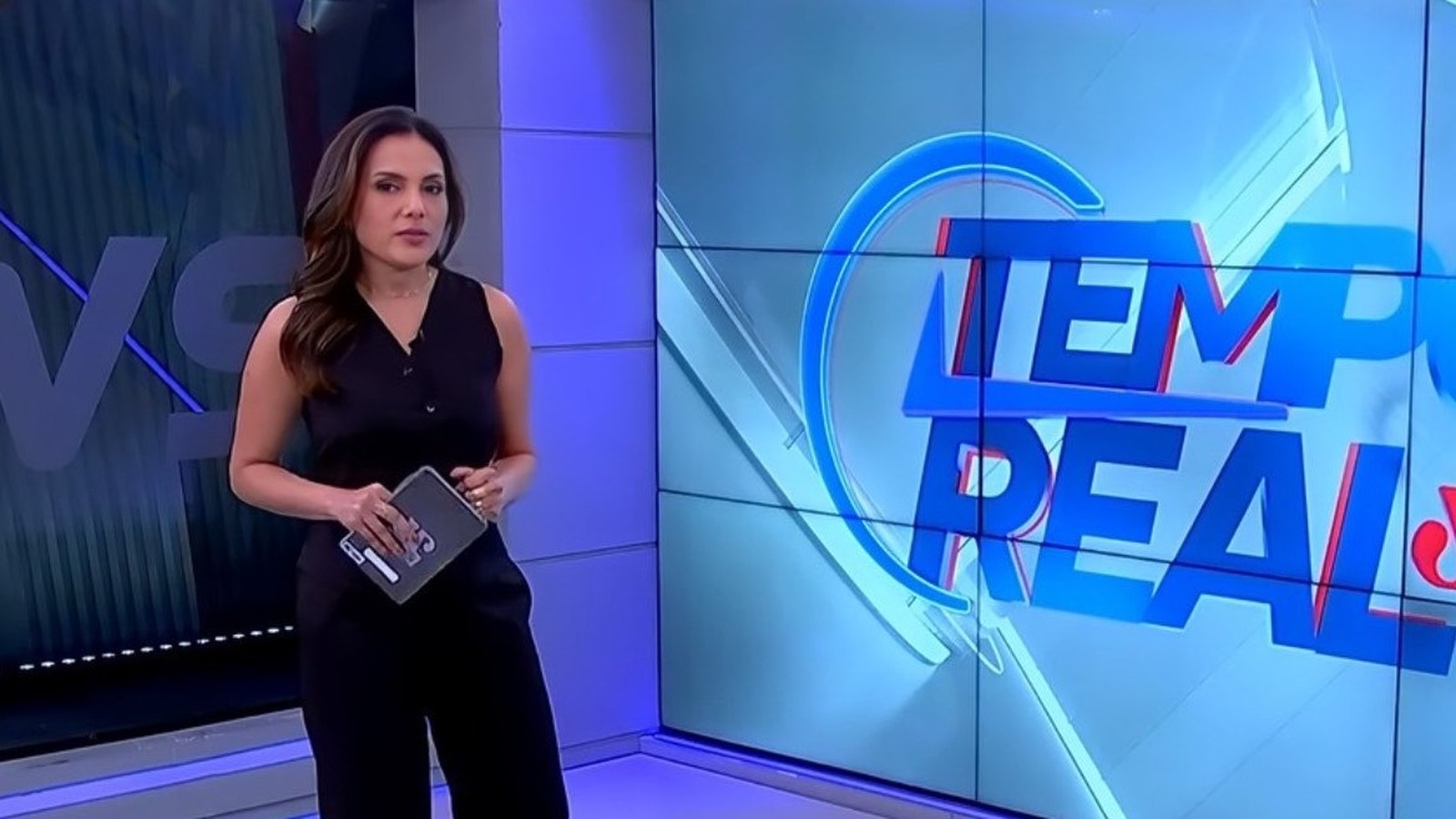 Apresentadora Márcia Dantas em estúdio, roupa preta, tablet nas mãos, tela "Tempo Real" ao fundo