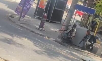 Dois homens em coletes táticos e fuzis, agachados atrás de postes em rua urbana