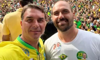 Dois homens sorrindo. Um com camisa amarela, outro com broches 'Bolsonaro 22', em multidão.