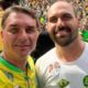 Dois homens sorrindo. Um com camisa amarela, outro com broches 'Bolsonaro 22', em multidão.