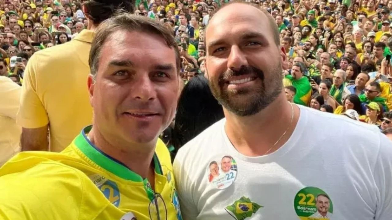 Dois homens sorrindo. Um com camisa amarela, outro com broches 'Bolsonaro 22', em multidão.