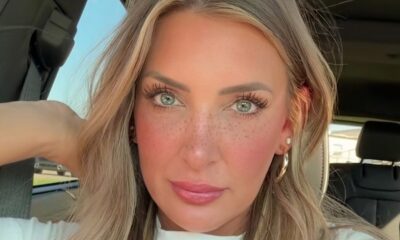 Close-up do rosto de uma mulher com cabelo loiro, olhos claros, sardas e maquiagem dentro de um carro