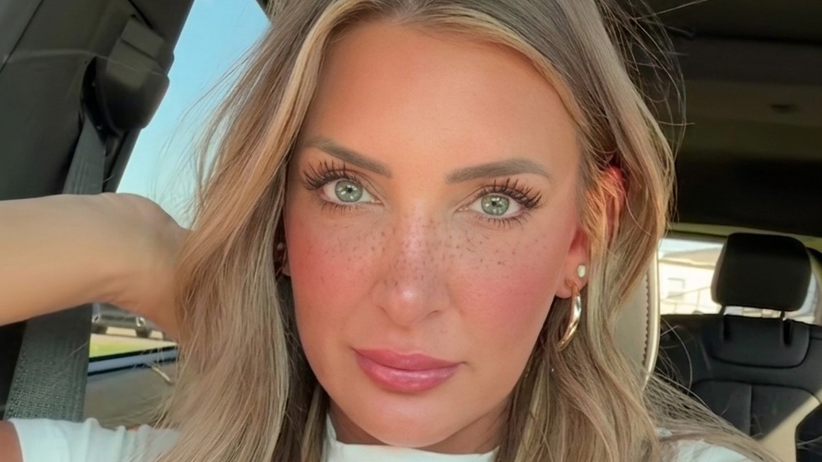 Close-up do rosto de uma mulher com cabelo loiro, olhos claros, sardas e maquiagem dentro de um carro