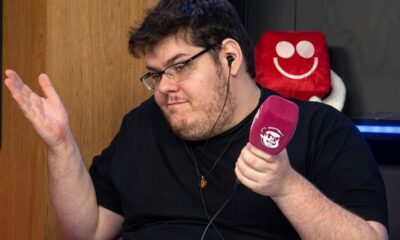 Homem de óculos, barba e fones de ouvido segura microfone roxo, gesticulando.