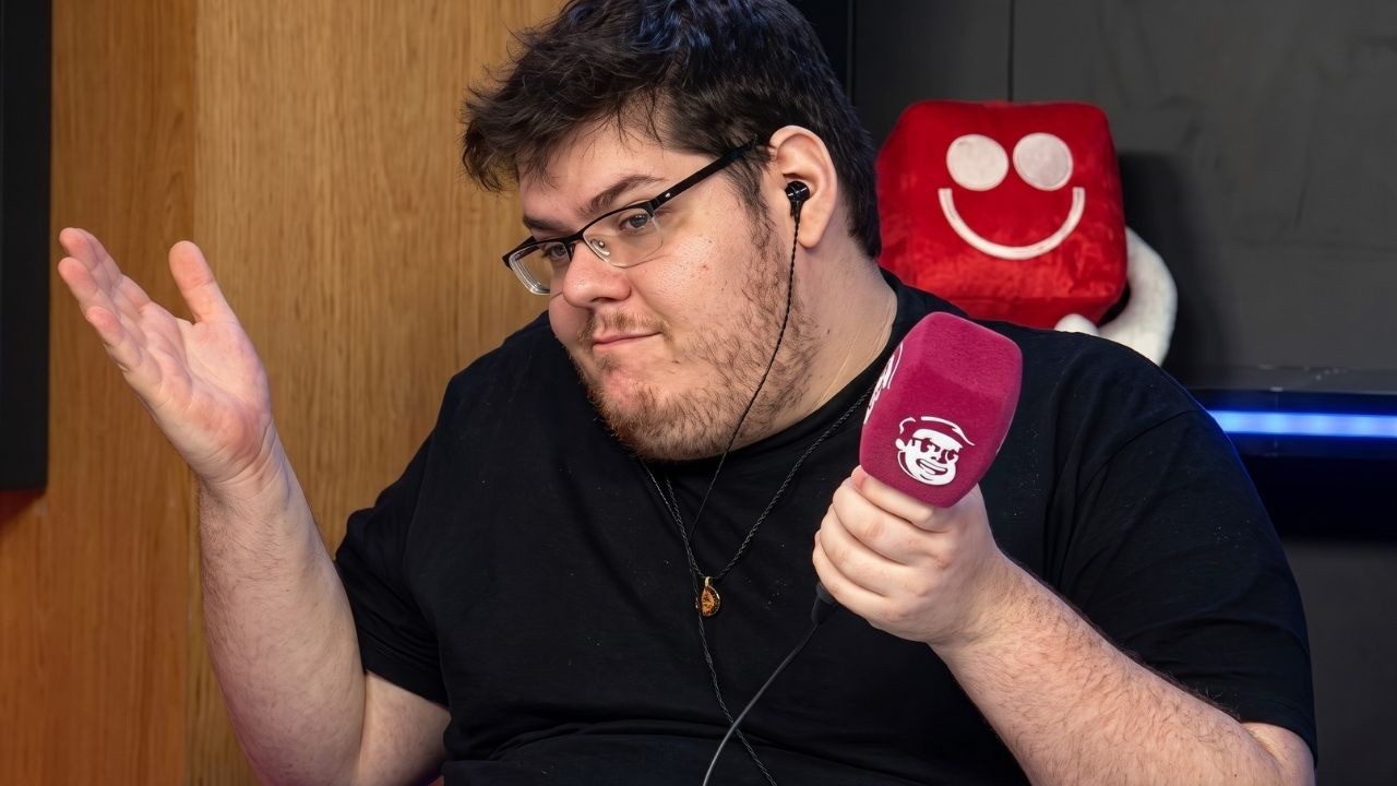 Homem de óculos, barba e fones de ouvido segura microfone roxo, gesticulando.