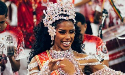 Mulher negra sorridente com coroa e fantasia brilhantes, faixa "Bellinha Delfim"