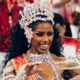 Mulher negra sorridente com coroa e fantasia brilhantes, faixa "Bellinha Delfim"