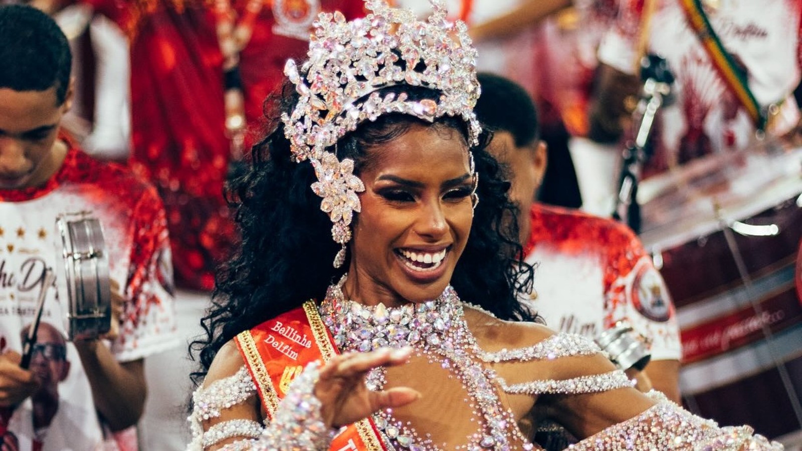 Mulher negra sorridente com coroa e fantasia brilhantes, faixa "Bellinha Delfim"