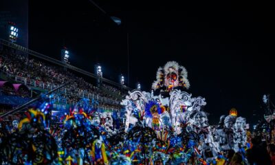 Detailed carnival float with feathered costumes, parade participants, and audience in stadium at night