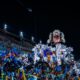 Detailed carnival float with feathered costumes, parade participants, and audience in stadium at night