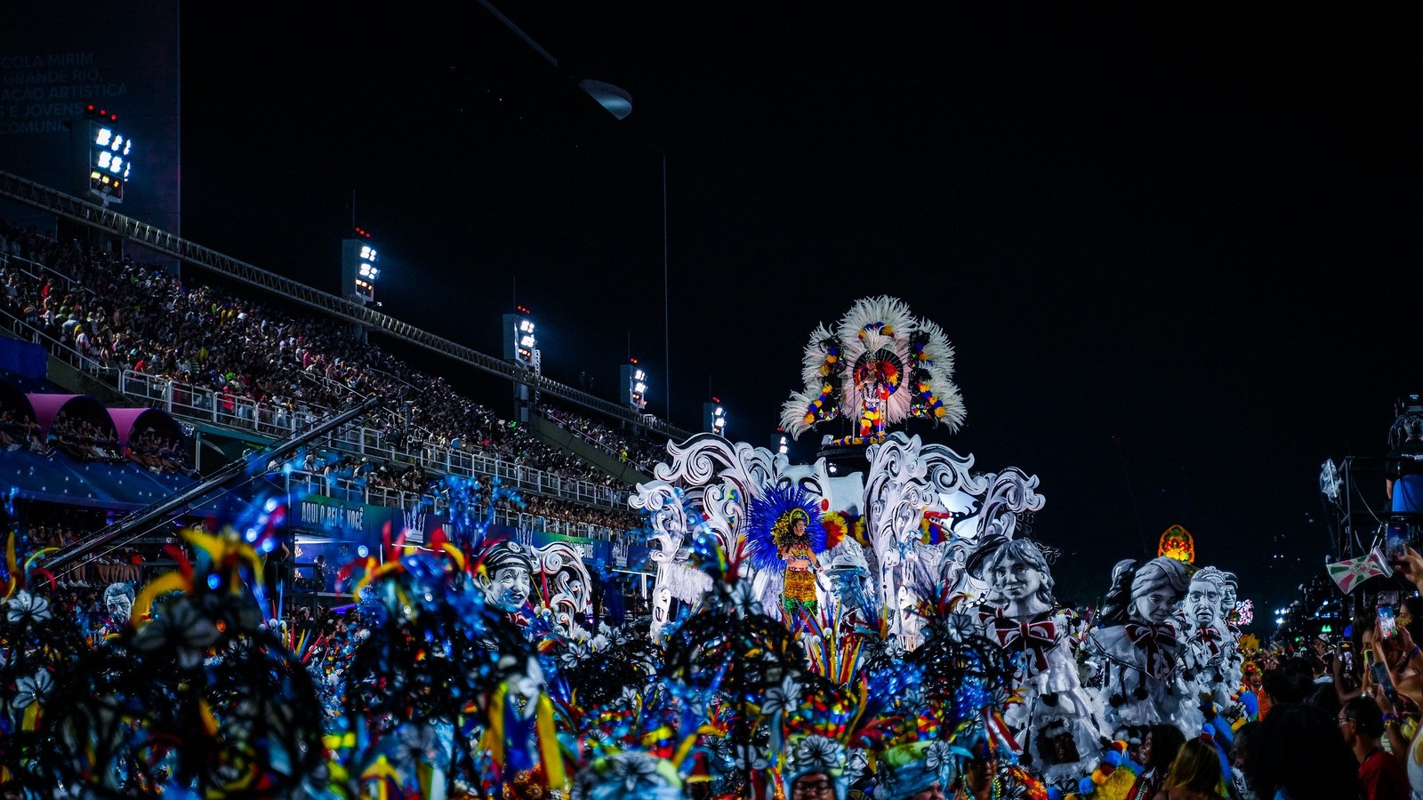Detailed carnival float with feathered costumes, parade participants, and audience in stadium at night