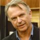 Retrato do ator Sam Neill, com cabelos grisalhos e camisa listrada, sorrindo levemente
