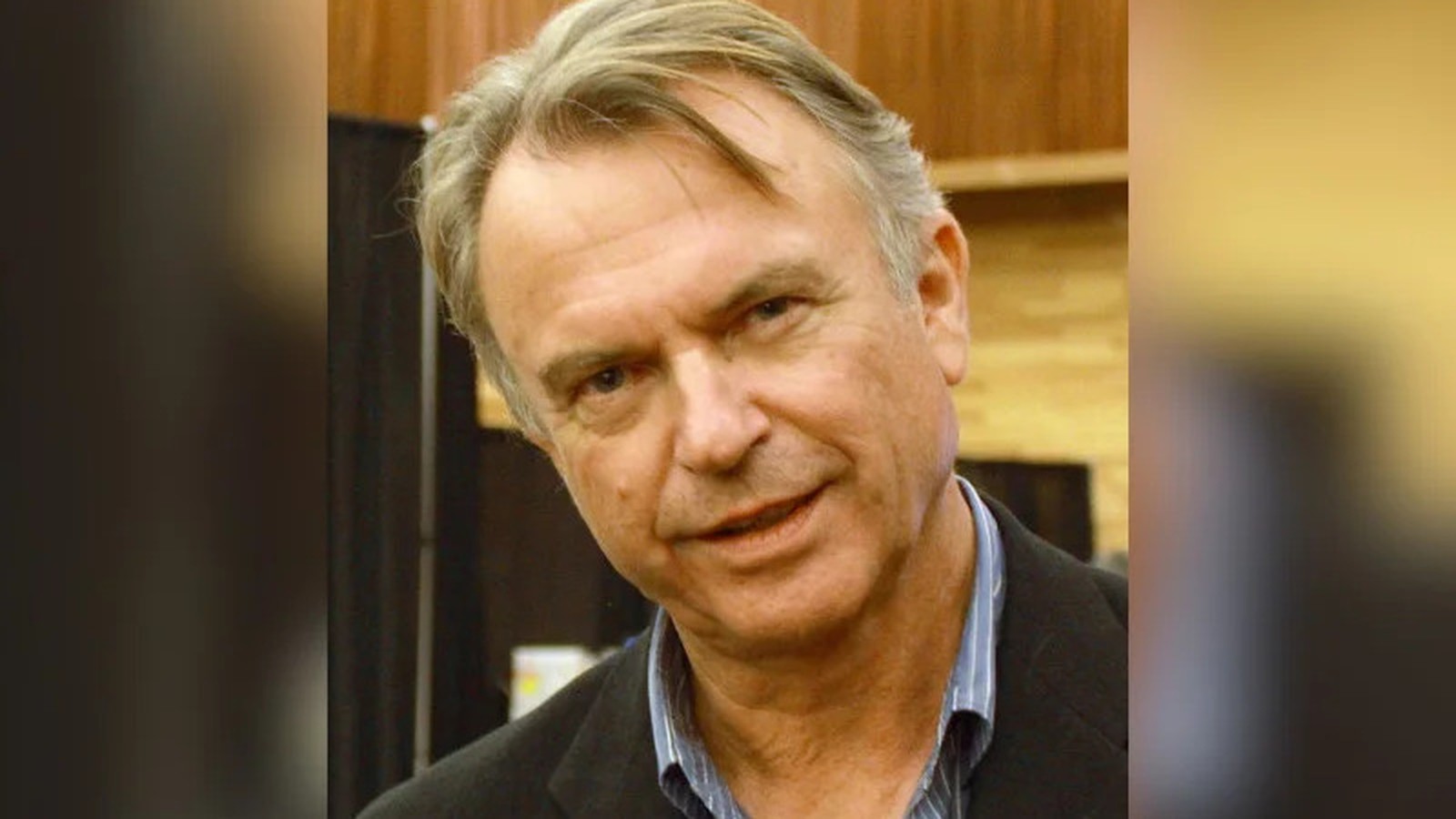 Retrato do ator Sam Neill, com cabelos grisalhos e camisa listrada, sorrindo levemente